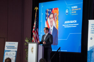 Jason Mahon on stage at a podium in front of a screen with his official portrait on it.